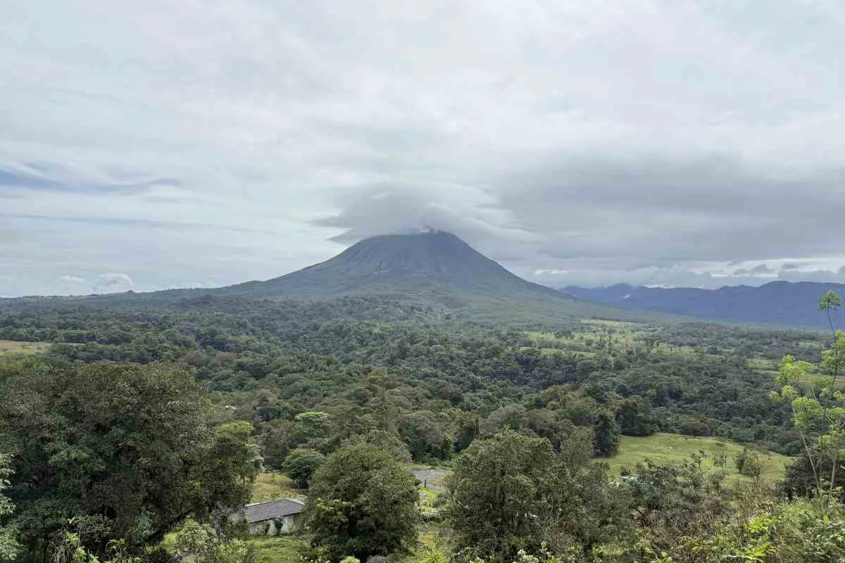 Blick über grünen Regenwald und Weiden auf den wolkenverhangenen Vulkan Arenal