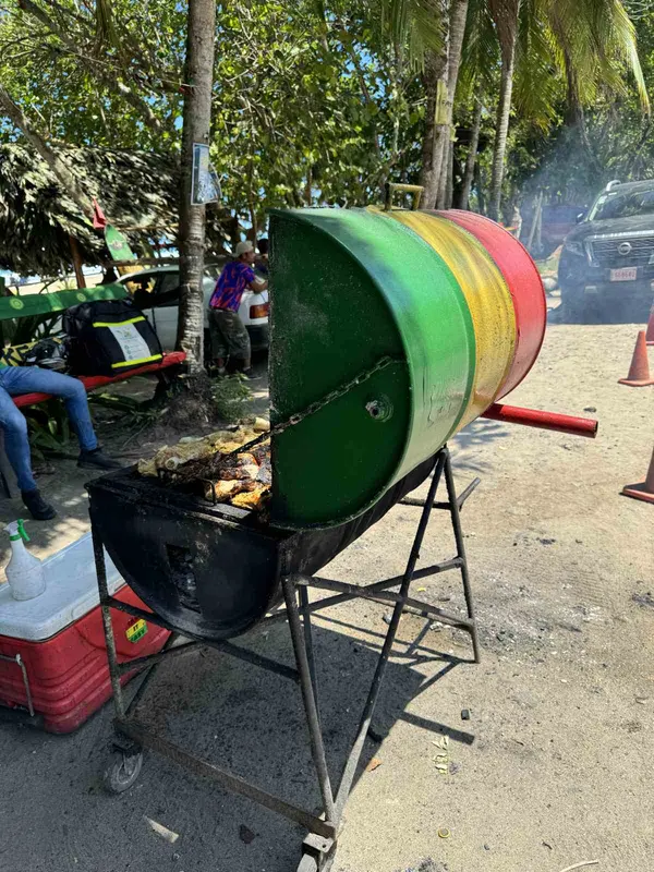 Grill aus einem rot-gelb-grün bemalten Fass am Straßenrand unter Palmen