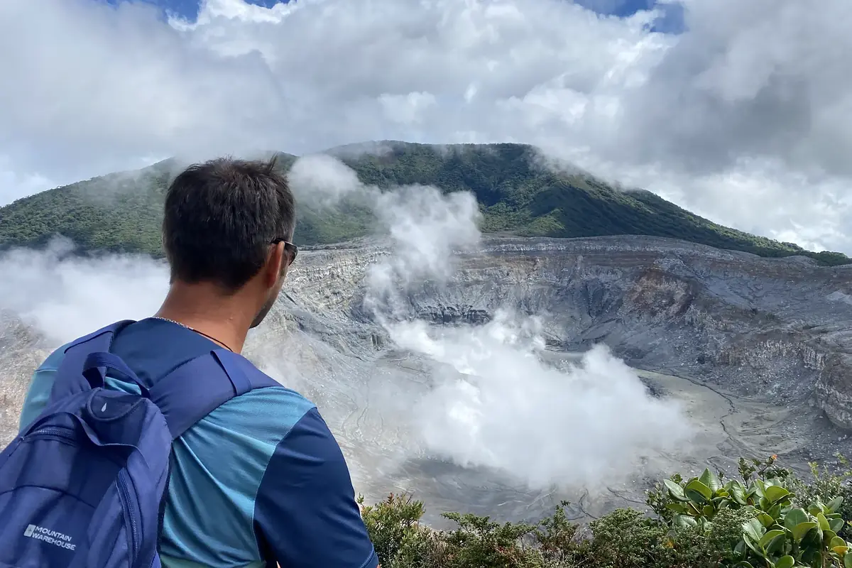 Blick über die Schulter eines Wanderers in den dampfenden Krater des Poás